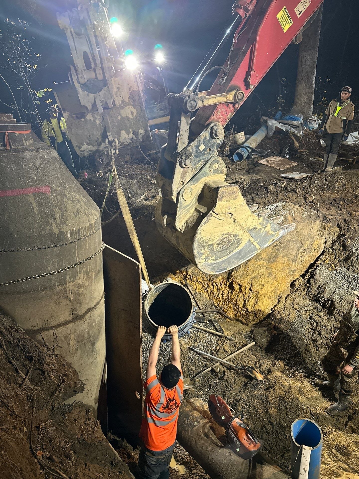 A crew guiding a lowered bucket load into a deep excavation under floodlight at night